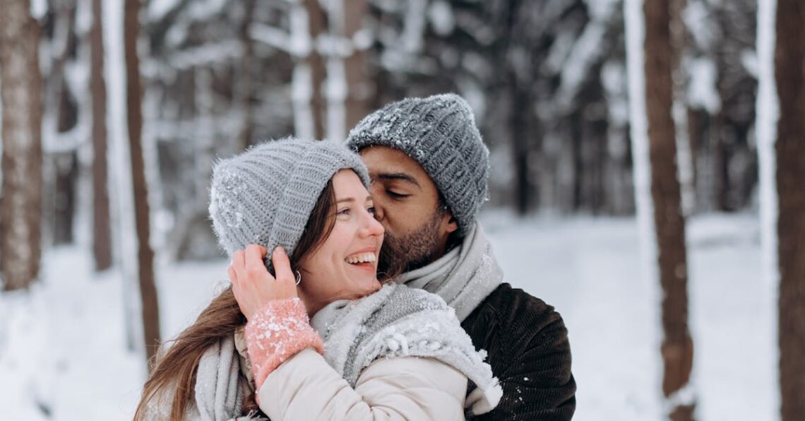 Couple embracing in winter attire amidst a snowy forest backdrop, radiating warmth and joy.