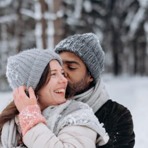 Couple embracing in winter attire amidst a snowy forest backdrop, radiating warmth and joy.