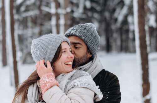 Couple embracing in winter attire amidst a snowy forest backdrop, radiating warmth and joy.